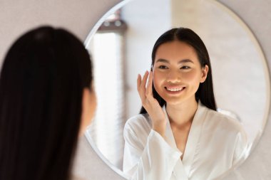 Bin yıllık ipek bornozlu güzel bir Koreli kadın nemlendirici yüz kremi sürüyor. Aynadaki yansımasına gülümsüyor, sabah cilt bakımı ritüelinin tadını çıkarıyor..