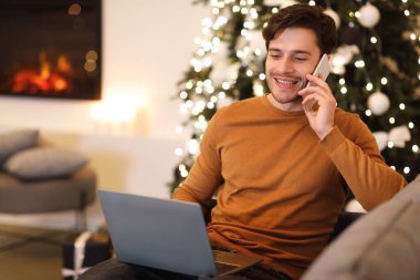 Bir adam dekore edilmiş bir oturma odasındaki koltukta rahat rahat oturup telefonuyla sohbet ediyor. Önünde önü açık bir dizüstü bilgisayar var, arkasında da festival ışıkları var..