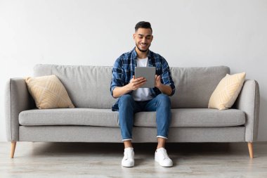 Gülümseyen genç bir Arap, dijital tablet kullanarak oturma odasındaki kanepeye rahatça oturur. İnterneti keşfetmekten ve eğlence seçeneklerini seçmekten mutlu..