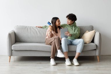 Young Asian couple sits on a gray sofa at home, using a tablet and a credit card to order goods online. They appear happy and engaged in their shopping experience.
