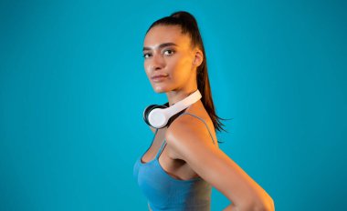 Spor elbiseli kadın, mavi bir arka planda kablosuz kulaklıkla rahatça poz veriyor. Onun güçlü varlığı ve odaklanması motivasyonu ve zindelik ruhunu ön plana çıkarıyor..