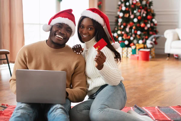 Evde Noel alışverişi için dizüstü bilgisayar kullanırken yerde oturan gülümseyen siyah çift. Kırmızı bir kredi kartı tutuyorlar, Noel Baba şapkaları takıyorlar, etrafı Noel süsleriyle çevrili..