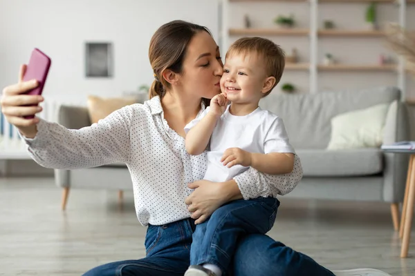 Bir anne, genç oğluyla yerde oturur ve neşeli bir selfie çeker. Oturma odasında yumuşak ışıklandırmalı ve modern dekorlu rahat bir atmosfer var..