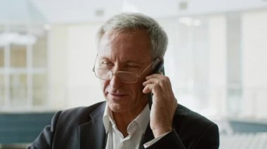 Takım elbiseli yaşlı bir adam modern bir ofiste telefonla konuşuyor. İşine odaklanmış ve profesyonel görünüyor. İş günlerinde kurumsal bir ortamda iş konularını tartışıyor..