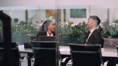 İş kıyafeti giymiş iki genç profesyonel modern bir şirket ofisinde dinamik bir sohbetin tadını çıkarıyorlar. Yeşilliklerle çevrili şık bir konferans masasında oturuyorlar..