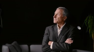 Terzi kıyafeti giymiş yaşlı bir adam kollarını kavuşturup çağdaş bir ofis ortamında yukarıya bakıyor. Atmosfer, liderliğe odaklanmış kurumsal bir yaşam tarzını yansıtıyor..