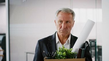 Yaşlı bir adam modern bir ofiste elinde kişisel eşyalarla dolu bir kutu tutuyor. Düşünceli görünüyor, yılların adanmışlığını ve kurumsal kariyerindeki sıkı çalışmayı yansıtıyor..