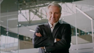 İş elbisesi içinde başarılı bir kıdemli adam modern ofis ortamında kollarını kavuşturmuş duruyor. Gösterişli mimari tasarımlar ve doğal ışık altında güven ve profesyonellik yayar..