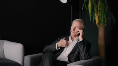 Takım elbiseli kıdemli bir işadamı, çağdaş bir ofiste otururken telefonla konuşuyor. Kendinden emin ve kurumsal bir konuşmayla meşgul görünüyor..