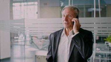 Takım elbise giymiş yaşlı bir adam modern bir ofiste duruyor ve bir telefon görüşmesi yapıyor. Etrafı kurumsal bir ortamla çevriliyken odaklanmış görünüyor, profesyonel bir yaşam tarzını yansıtıyor..