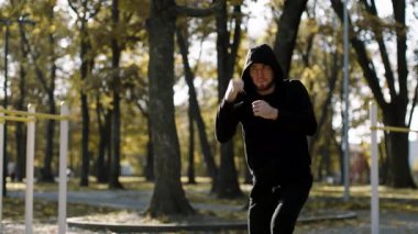 A young man dressed in a black hoodie practices fitness moves in a park surrounded by autumn foliage. He focuses on his routine in the vibrant outdoor setting, embodying a healthy lifestyle.