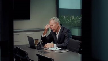 Takım elbiseli kıdemli bir adam ofisteki konferans masasında oturuyor. Alnını eline dayar, laptopuyla çalışırken ezilmiş gibi görünür, iş malzemeleriyle çevrilidir..