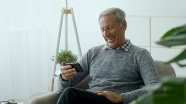 60 'lı yaşlarda bir adam rahat bir şekilde oturma odasında otururken akıllı telefonuyla meşgul olmayı sever. Güneş ışığı pencereden süzülüp sıcak bir atmosfer yaratır..