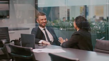 İki genç profesyonel modern bir şirket ofisinde stratejileri tartışıyor. Atmosfer işbirlikçi, yeni nesiller arasındaki ticarete dinamik bir yaklaşımı vurguluyor..
