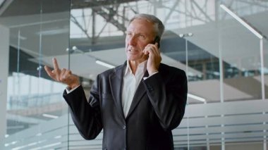 İş kıyafeti giymiş yaşlı bir adam düşünceli bir şekilde el kol hareketi yaparken bir telefon görüşmesi yapar. Çağdaş bir ofiste duruyor, kurumsal yaşam tarzını ve liderlik rolünü yansıtıyor..