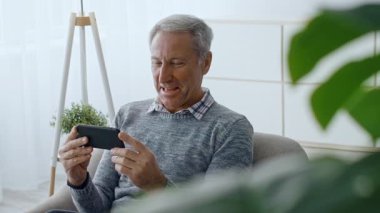 60 'lı yaşlarda yaşlı bir adam evinde rahat bir şekilde oturuyor, akıllı telefonuyla meşgul. Etkileşim kurarken gülümsüyor, rahat bir yaşam tarzındaki keyif anlarını vurguluyor..