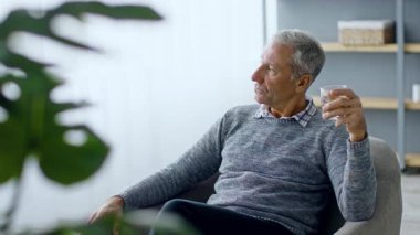 60 'lı yaşlarda bir adam evinde rahat bir sandalyede oturup içkisini yudumlarken dikkatlice seyrediyor. Atmosfer, modern ortamda huzurlu bir yaşam tarzını yansıtıyor..