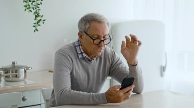 60 'lı yaşlarda bir adam mutfak masasında oturuyor ve akıllı telefonuna odaklanıyor. Gözlük takıyor ve evde rahat bir yaşam tarzı yayarak günlük aktivitelerle meşgul..