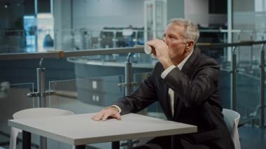 Resmi takım elbiseli yaşlı bir adam kurumsal bir ofiste bir masada oturuyor, bir fincandan yudumluyor. Çevresindeki modern çalışma alanı, kurumsal yaşam tarzını vurgulayan profesyonel bir atmosfer sergiliyor..