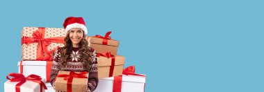 Noel Baba şapkası takan ve elinde hediye tutarken gülümseyen bir kadın. Arkasında güzel paketlenmiş kurdeleli hediyeler var. Bayram kutlamalarının neşeli ruhunu yakalıyorlar..