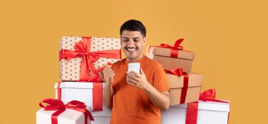 Bir adam renkli hediye kutularıyla çevrili akıllı telefonuna bakarken geniş bir gülümseme sergiliyor. Neşeli atmosfer heyecan ve mutlulukla dolu şenlikli bir olayı akla getiriyor..