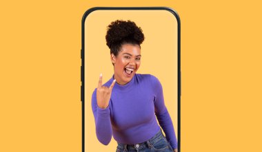 Genç bir kadın akıllı telefon çerçevesi içinde hevesli bir el hareketi göstererek mutlu bir şekilde poz veriyor. Mor bir bluz giyiyor ve canlı sarı bir arka plana gülümsüyor, canlı bir anı yakalıyor..