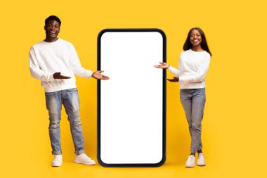 Genç bir çift, her biri büyük boş bir akıllı telefona doğru işaret eden, canlı sarı bir arka planda güvenle duruyor. İfadeleri neşeli, davetkar bir atmosfer yaratıyorlar..