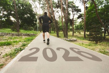 Bir kişi 2026 sayısıyla işaretlenmiş yemyeşil bir parkta asfalt bir yolda koşuyor. Ağaçlar ve doğa, yeni bir başlangıç ve kararlılığa işaret eden sakin bir atmosfer yaratırlar..