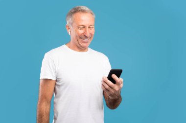 Gülümseyen olgun bir adam mavi arka planda duruyor, cep telefonunu kullanıyor ve kablosuz kulaklık takıyor. İnternette sohbet ederken, müzik dinlerken ve anın tadını çıkarırken mutlu görünüyor..