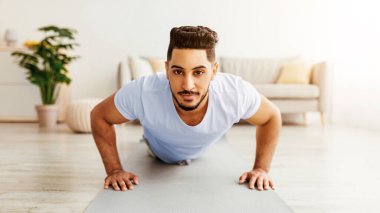 Bir adam, iyi aydınlatılmış bir oturma odasında yoga minderinde şınav çekiyor. Bu alan modern ve rahat bir tasarım içinde bitki ve mobilya barındırıyor..