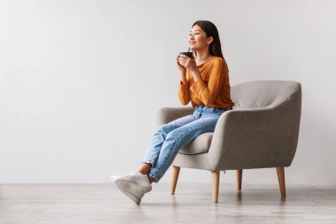 Güzel genç bir Asyalı kadın rahat bir koltukta dinlenirken kahvesini yudumluyor. Parlak beyaz bir duvara karşı mola veriyor, huzurlu bir anın tadını çıkarıyor..