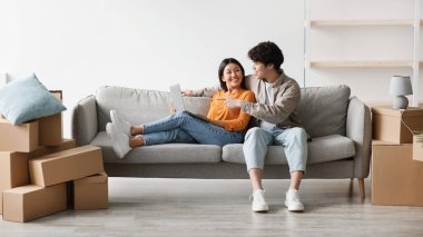 Happy millennial Asian couple sits comfortably on a couch among cardboard boxes as they browse the internet for home decoration ideas and plan their new living space together.