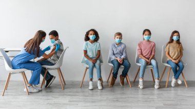 Çocuklar sabırla Covid-19 aşılarını yöneten kadın hemşire olarak otururlar. Çeşitli gruplar, tarafsız bir geçmişe sahip çok ırklı erkek ve kızları içeriyor. Sağlıklarına öncelik veriliyor..