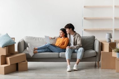 Cheerful young Asian couple sits on a couch browsing a laptop, surrounded by cardboard boxes. They are excitedly exploring home decor ideas for their new apartment in a bright living room.