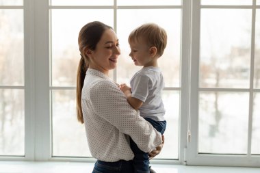 Bir anne oğlunu doğal ışıkla dolu sıcak bir odada kucaklıyor. Karlar pencerelerin dışına nazikçe düşerken birbirlerine gülümseyerek neşeli bir an yaşarlar..