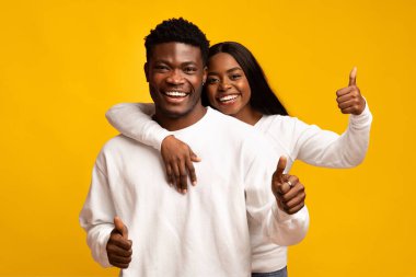 Neşeli bir çift mutlu bir anı paylaşır, birlikte poz verir, gülümser ve başparmak kaldırırlar. Günlük beyaz kıyafetleri, parlak sarı arkaplanla çok güzel tezat oluşturuyor..