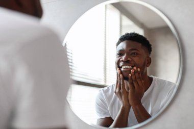 Neşeli bir Afrikalı Amerikalı adam banyoda yuvarlak bir aynanın önünde duruyor. Tıraşsız çenesine dokunurken yansımasına gülümsüyor, evde sabah bakım rutininin keyfini çıkarıyor..