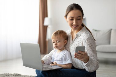 Parlak bir oturma odasında, bir anne yerde oturur, küçük çocuğu dizüstü bilgisayarla oynarken akıllı telefonuna odaklanır. Sıcak ve davetkar bir atmosferle çevrilidirler..
