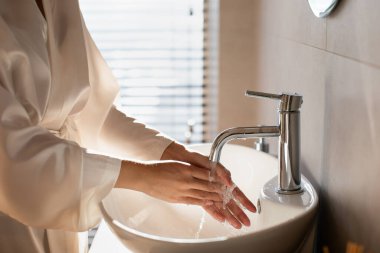 Beyaz ipek bornozlu bir kadın şık bir banyoda ellerini sabunla yıkıyor. Bu yakın çekim, hijyen ve kendine bakmanın önemini vurguluyor..