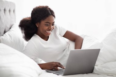 Neşeli, uzun saçlı kıvırcık saçlı, Afro-Amerikan milenyum kadını laptopunu kullanırken yatakta dinleniyor. Sosyal medyayı ya da interneti kontrol ediyor, pijamalarıyla rahat bir hafta sonu sabahının tadını çıkarıyor..