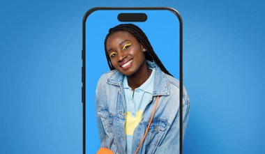 Örgülü genç bir kadın, kulaklıkla poz verirken kot ceket ve parlak makyaj yapar. Kendine güvenerek gülümsüyor, akıllı telefon tarafından çerçevelenmiş mavi bir arkaplana karşı..