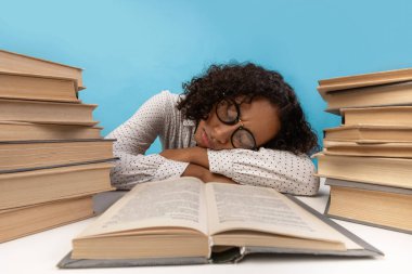 Yorgun bir Afrikalı Amerikalı kız öğrenci, kitap yığınları arasında başını masasında dinlendiriyor. O, mavi bir arkaplan karşısında yaklaşan sınavı için çalışırken bunalmış görünüyor..