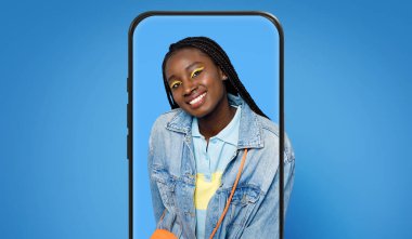 Örgülü ve parlak sarı makyajlı bir kız akıllı telefon çerçevesinde poz verirken mutlu bir şekilde gülümsüyor. Kot ceket giyiyor ve neşeli renkler giyiyor, neşeli bir atmosfer yaratıyor..