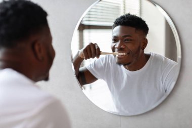 Afrikalı Amerikalı adam aynaya bakarken dişlerini fırçalar. Ağız temizliğine önem verirken gülümsüyor. Dekor, çağdaş özellikleri olan şık bir banyo..