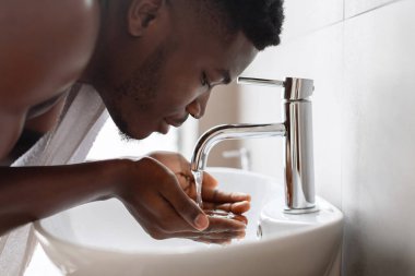 Afro-Amerikalı bir adam şık bir banyodaki bir lavaboda üstsüz duruyor, sabahın erken saatlerinde yüzünü suyla yıkıyor..