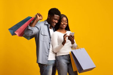 Genç bir çift, ellerinde alışveriş torbalarıyla parlak sarı bir arka plana karşı duruyor ve akıllı telefona bakıyor. Gülümsüyorlar ve alışveriş yaparken keyifli bir anı paylaşıyorlar..