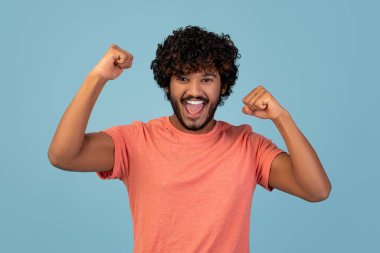 Kıvırcık saçlı ve sakallı yakışıklı bir Hintli kutlama için elini kaldırıyor. Mavi arka planda pembe bir tişört giyiyor. Heyecan ve başarıyı ifade ediyor..