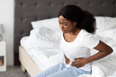 Genç bir Afro-Amerikan kadın sabah yatağında oturuyor, karın ağrısından açıkça etkilenmiş. Göbeğine dokunur ve regl olmanın verdiği rahatsızlığı gösteren pijamalar giyer..