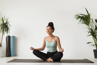 Sakin ve rahat esmer bir kadın evde yoga minderinin üzerinde nilüfer pozisyonunda oturur ve meditasyon yapar. Yumuşak ışık odayı dolduruyor. Etrafındaki yeşilliklerle huzurlu atmosferi güçlendiriyor..