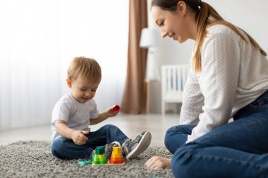 A young child engages with colorful stacking toys while seated on a soft rug. A smiling mother watches nearby, enjoying the playful interaction in a sunny living room.